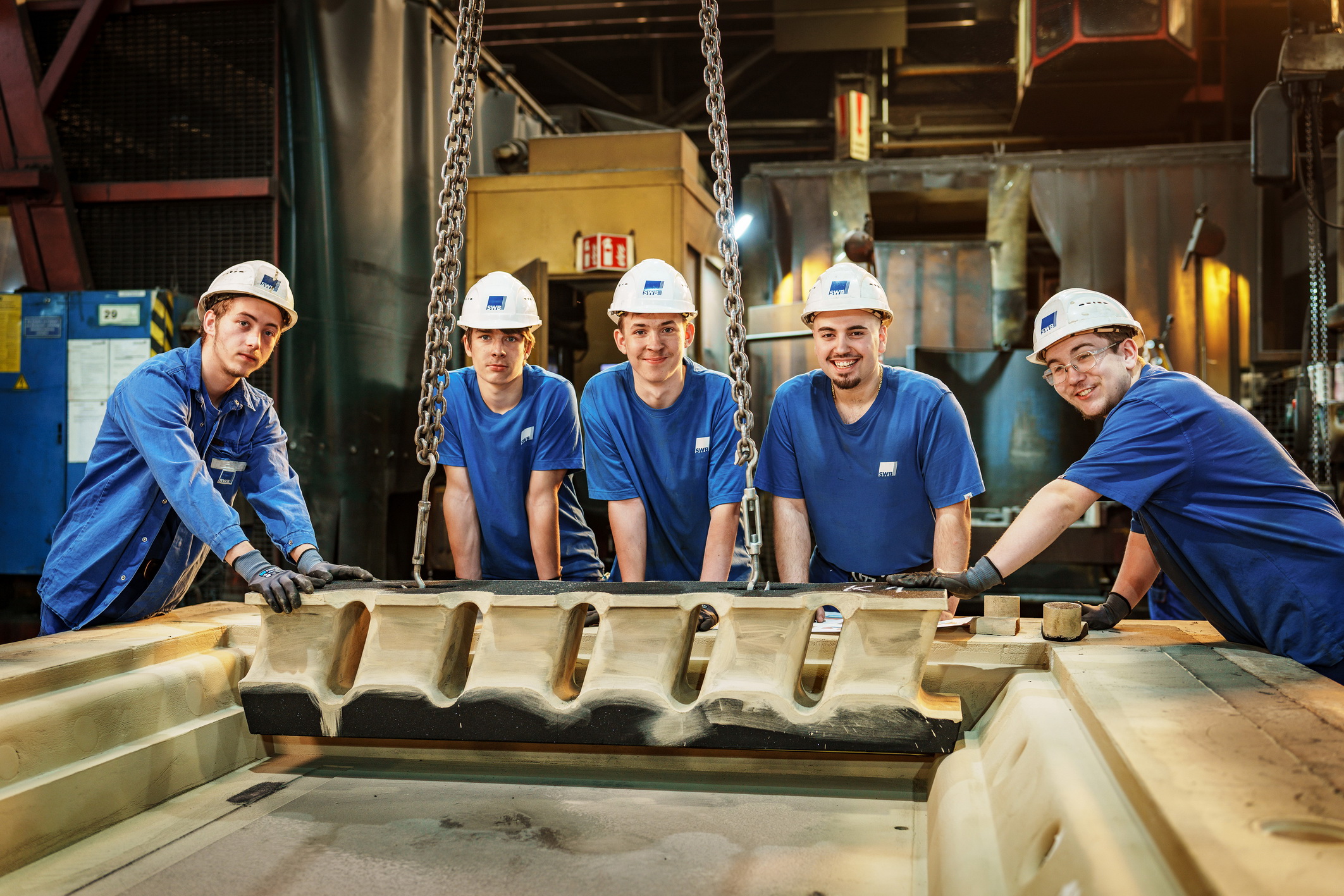 5 Auszubildende in blauen T-Shirts und mit weißem Helm mit SWB-Logo stehen an einer Gießform - Five apprentice workers wearing blue t-shirts and white helmets with the SWB logo are standing next to a casting mold